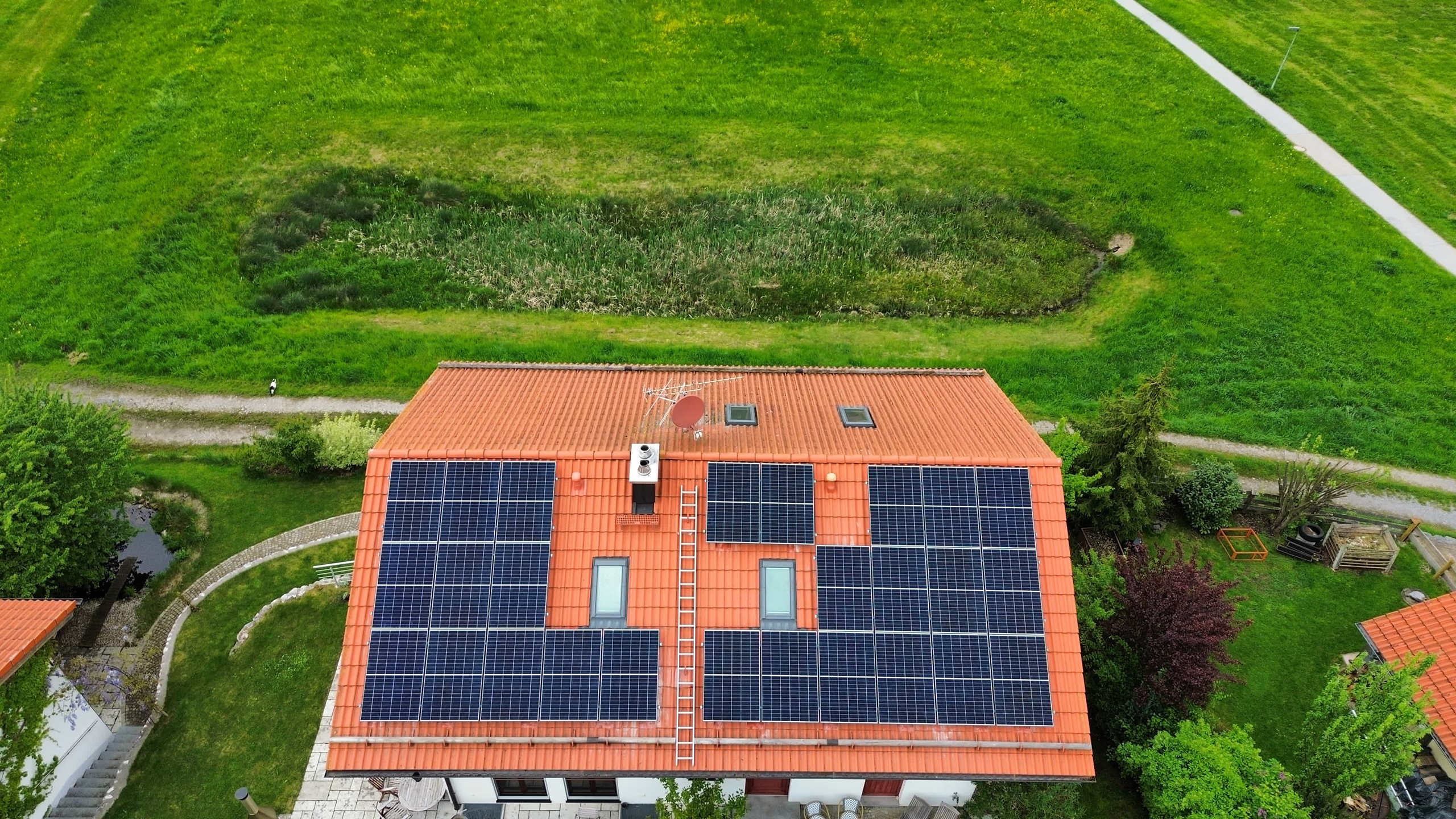 Eine private Solaranlage auf einem roten Ziegeldach. Das Haus steht inmitten eines großen grünen Grundstücks mit Wegen, Bäumen und Gartenflächen.