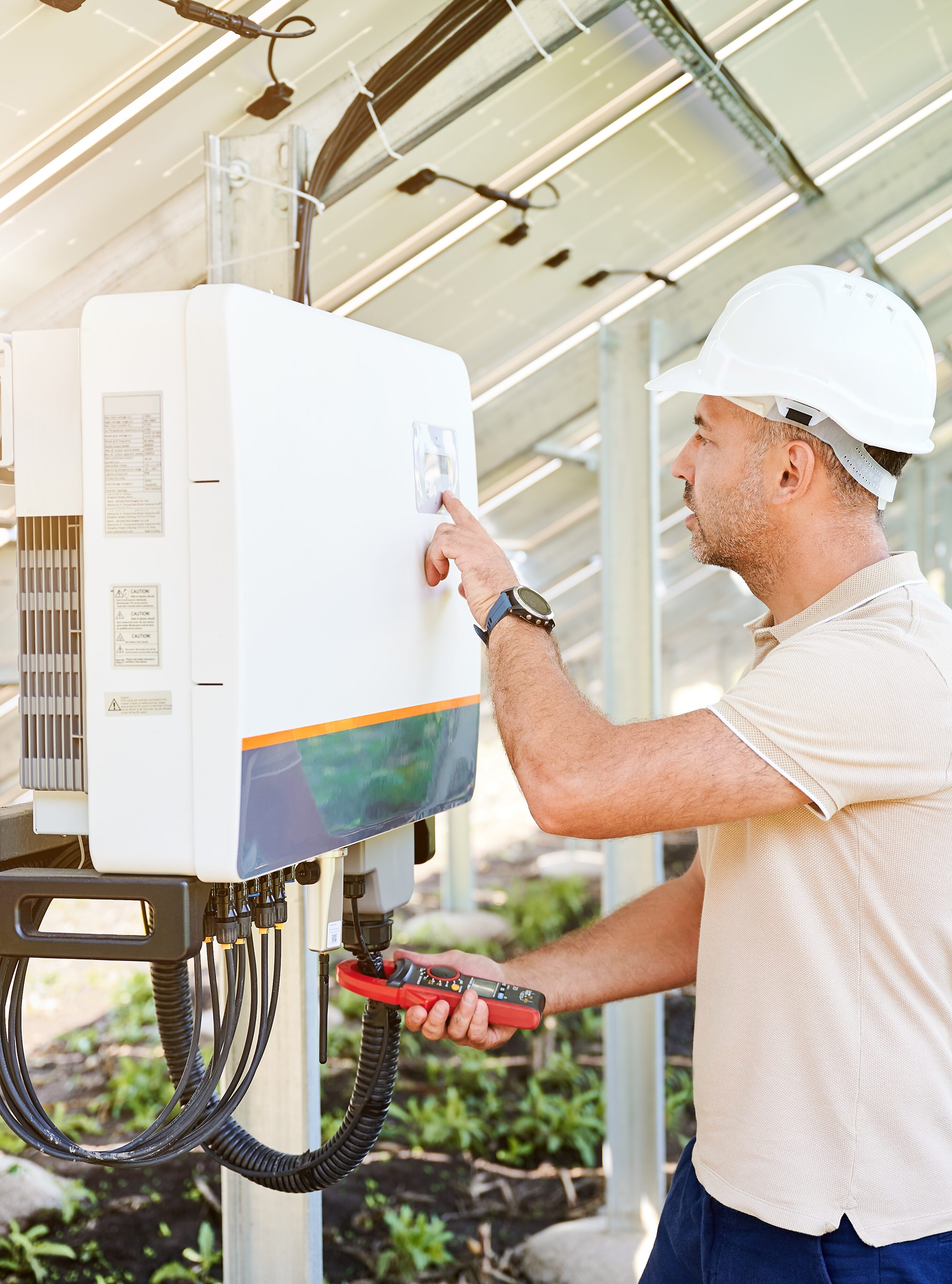 Ein Techniker überprüft einen Photovoltaik-Wechselrichter auf einer Freiflächenanlage. Er trägt einen weißen Schutzhelm und hält ein Messgerät in der Hand.